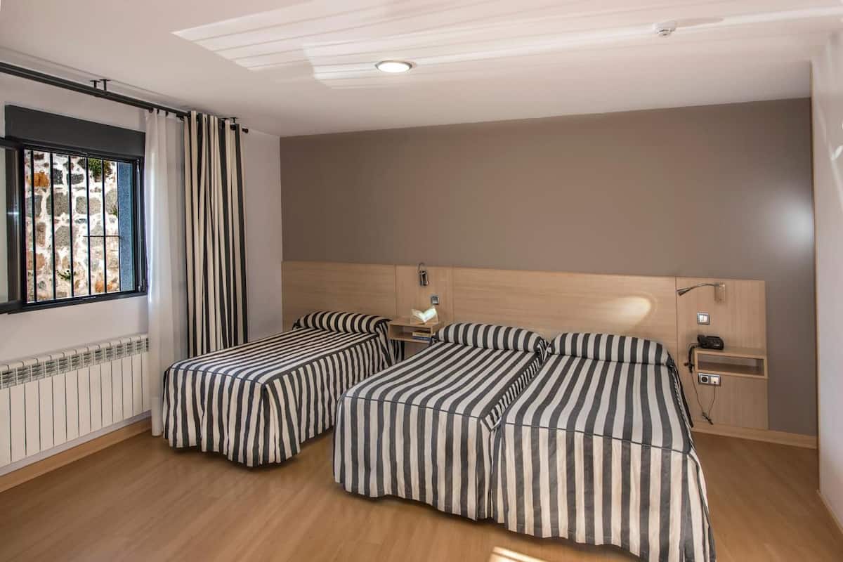 Une chambre avec deux lits, une fenêtre avec des rideaux, un radiateur et une tête de lit en bois.