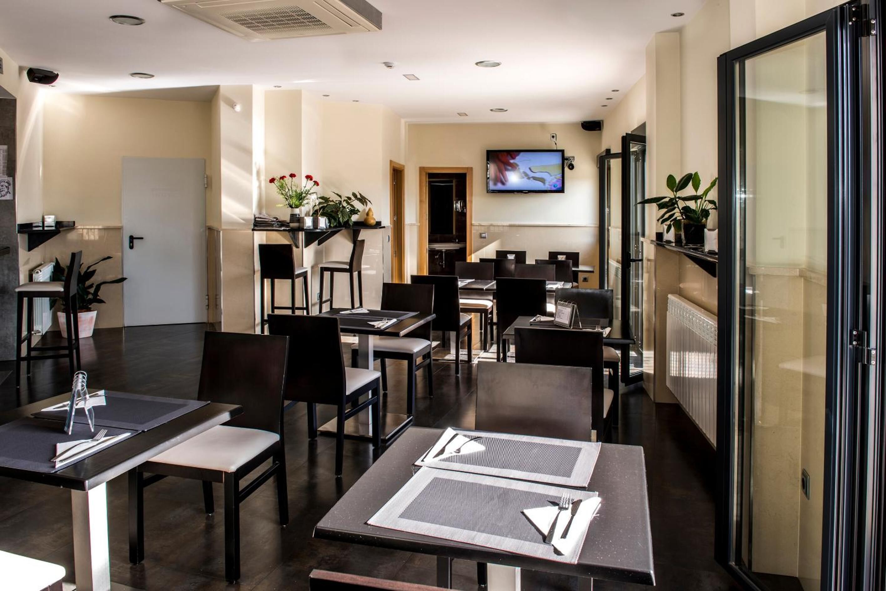 Una sala da pranzo di un hotel moderno, con tavoli apparecchiati per gli ospiti, una televisione a parete e una pianta in vaso sul davanzale.