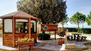 A wooden gazebo with a bench and a planter with flowers, surrounded by a gravel area and trees. - Terra dei Santi Country House (Spello)
