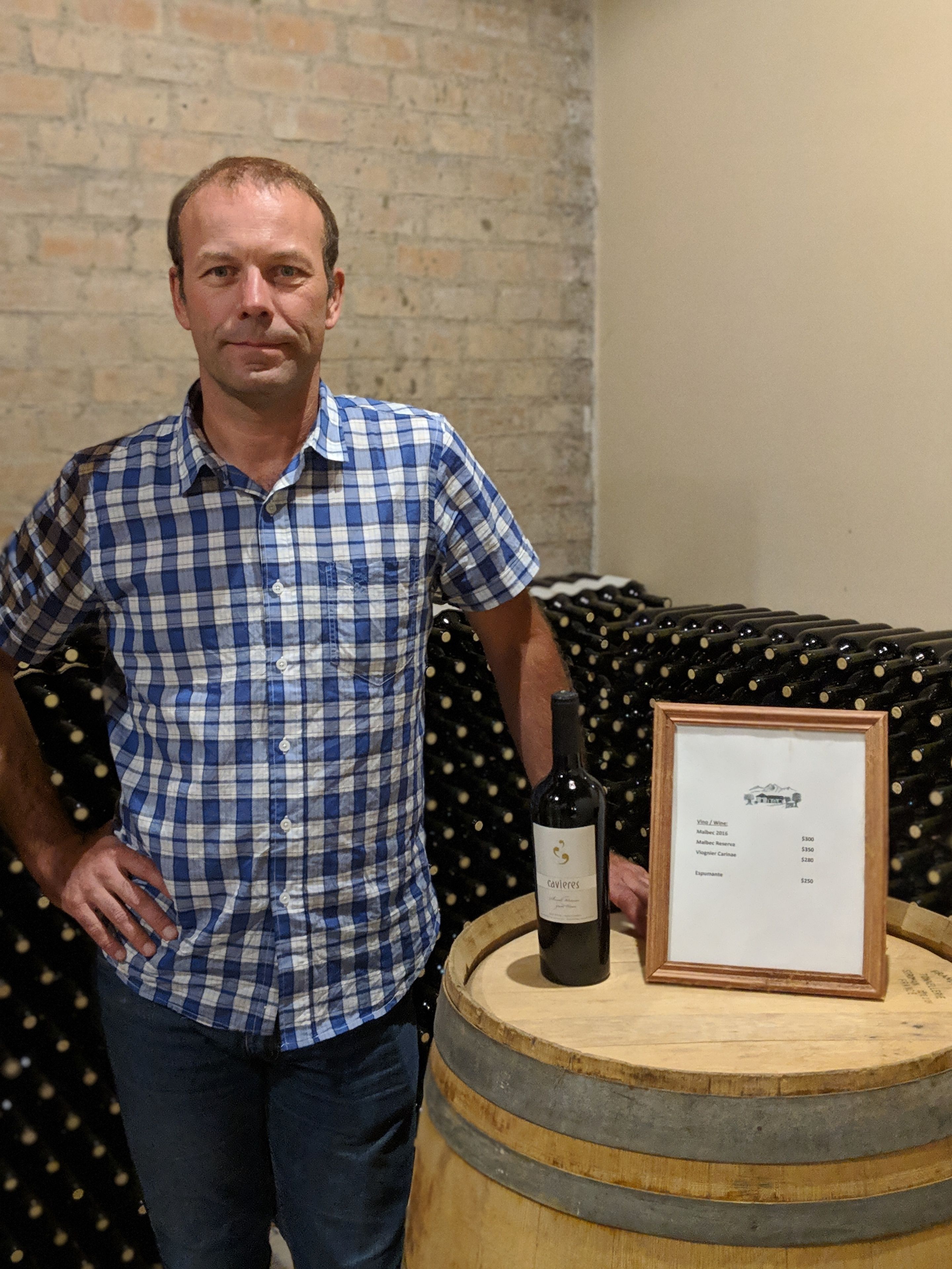 A man standing next to a wooden barrel with a wine bottle and a framed menu.