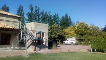 A house with a concrete staircase leading to a balcony, a white SUV parked in front, and a lush green garden.