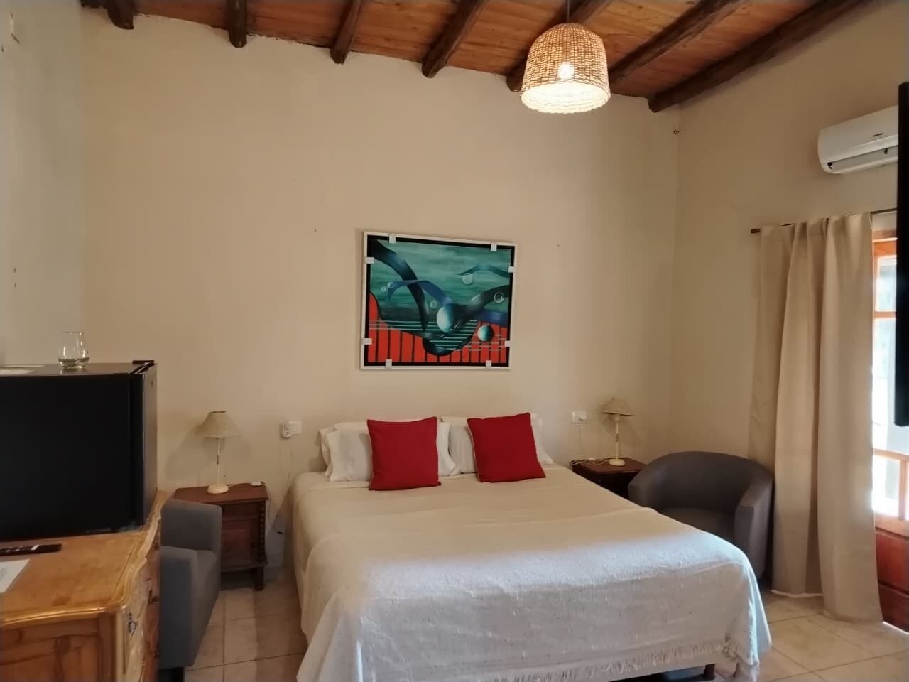 A bedroom with a bed, a desk, a chair, a lamp, a painting, and an air conditioning unit.