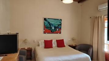 A bedroom with a bed, a desk, a chair, a lamp, a painting, and an air conditioning unit.