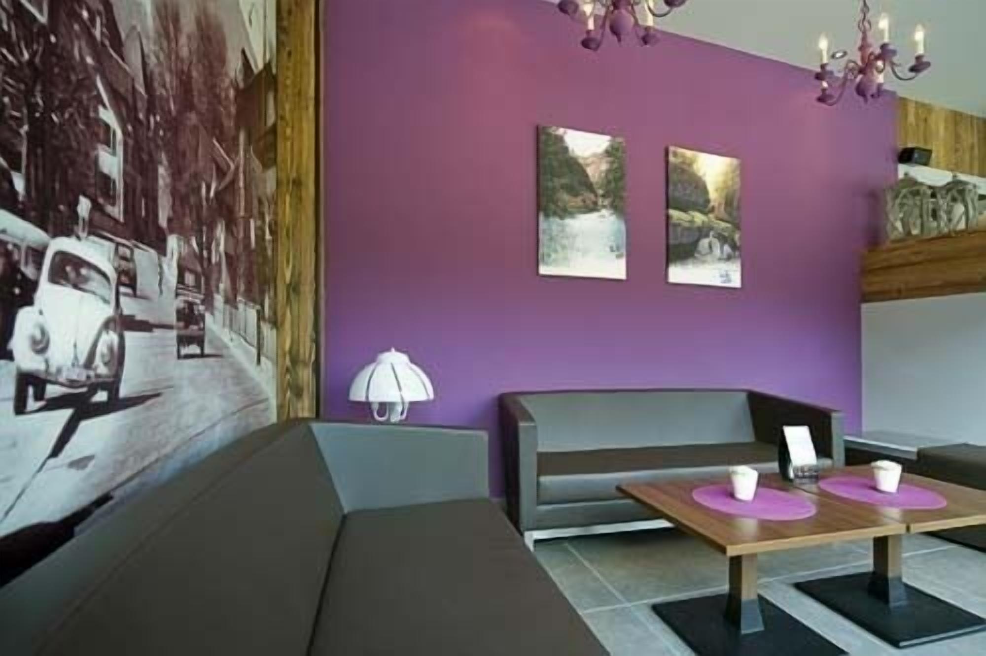 Un hall d’hôtel moderne avec un mur violet, un grand tableau représentant une voiture, un banc et une table avec deux tasses.