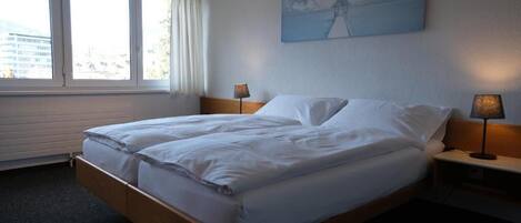 Una habitación de hotel con cama, mesita de noche, lámpara, ventana con cortinas y un cuadro en la pared.