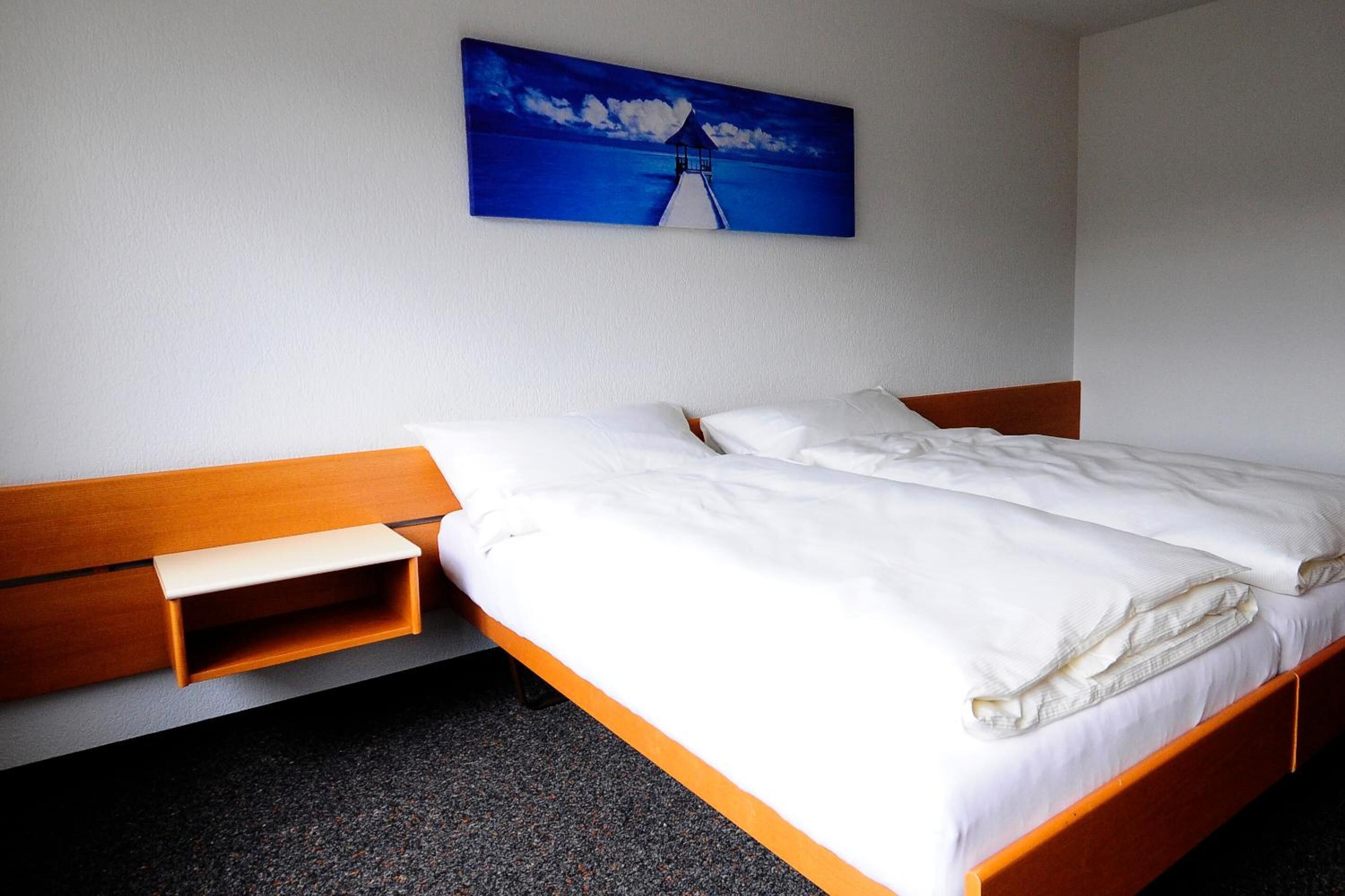 Ein Einzelbett mit hölzernem Kopfteil und Fußteil, ein Nachttisch sowie ein Gemälde eines Leuchtturms an der Wand.