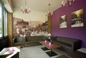A modern interior with a purple wall, a sofa, a table with cups, and a mural of a street scene. - Hôtel Oasis (Moutier)