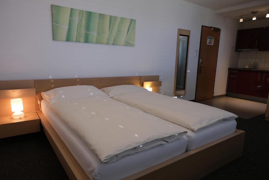 A double bed with a wooden headboard, bedside lamps, a mirror, and a kitchen area with cabinets.
