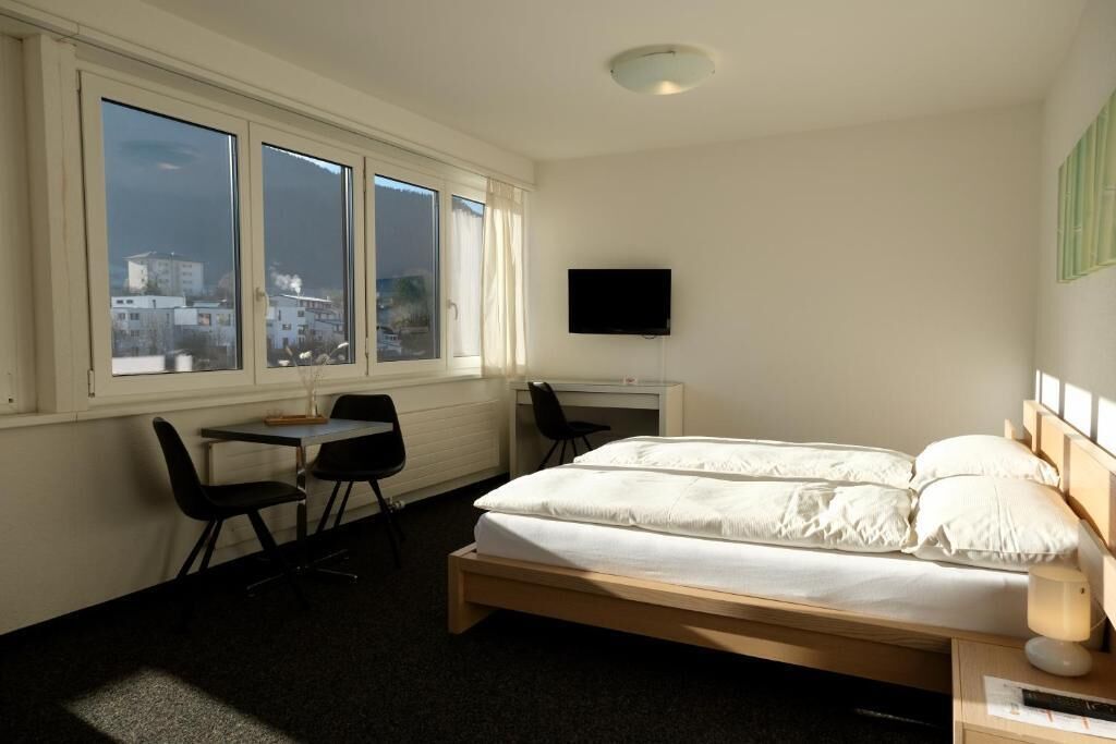 Une chambre à coucher comprenant un lit, un bureau, une chaise, un téléviseur et une fenêtre donnant sur la ville.