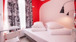 A hotel room with a bed, white pillows, a desk, a chair, and a window with patterned curtains. - La Guitarra Hostel (Gdansk)