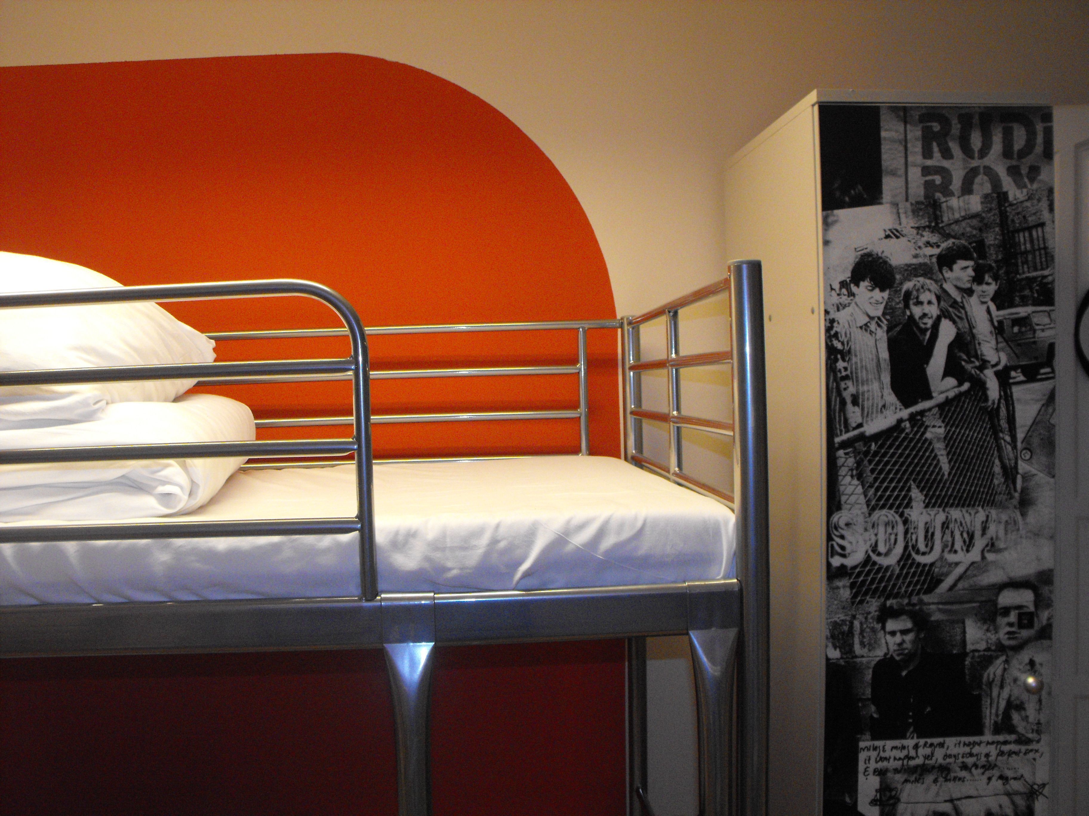 Een stapelbed met metalen spijlen en een witte matras, een rode muur en een poster met een band erop.