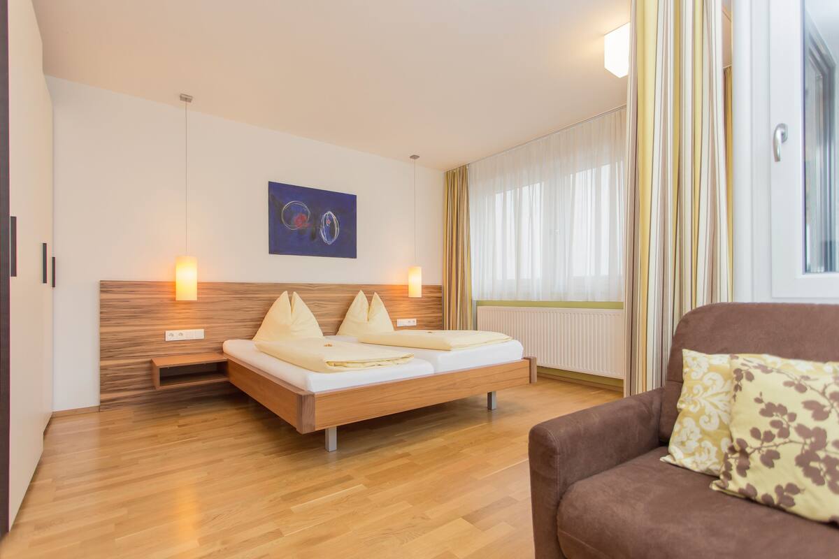 Una habitación de hotel moderna con una cama de madera, un sofá marrón y un gran ventanal con cortinas.