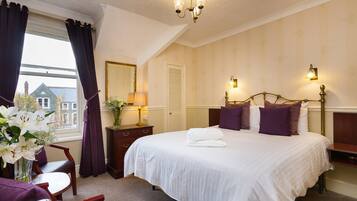A neatly arranged bedroom with a large bed, a bedside table, a chair, and a window with curtains.