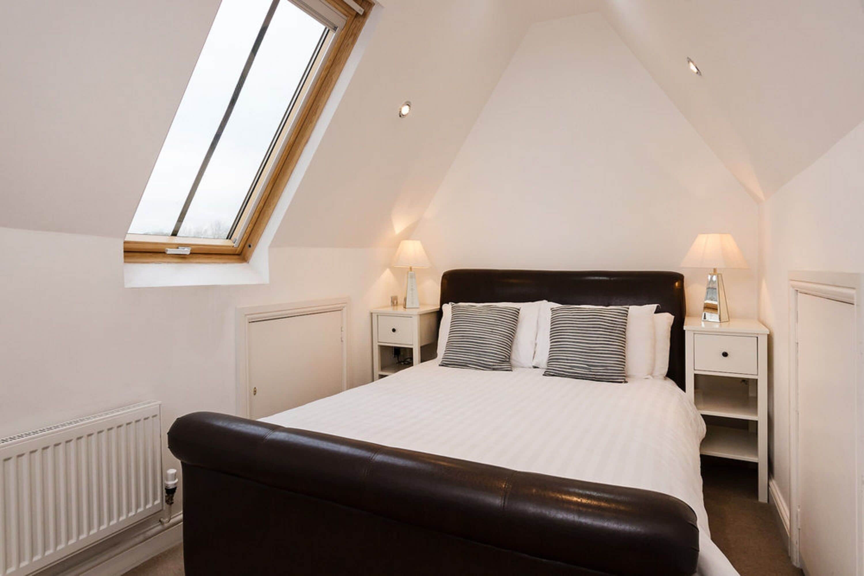 A bedroom with a sloped ceiling, a skylight, a large bed with striped pillows, two bedside tables with lamps, and a radiator.