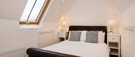 A bedroom with a sloped ceiling, a skylight, a large bed with striped pillows, two bedside tables with lamps, and a radiator.