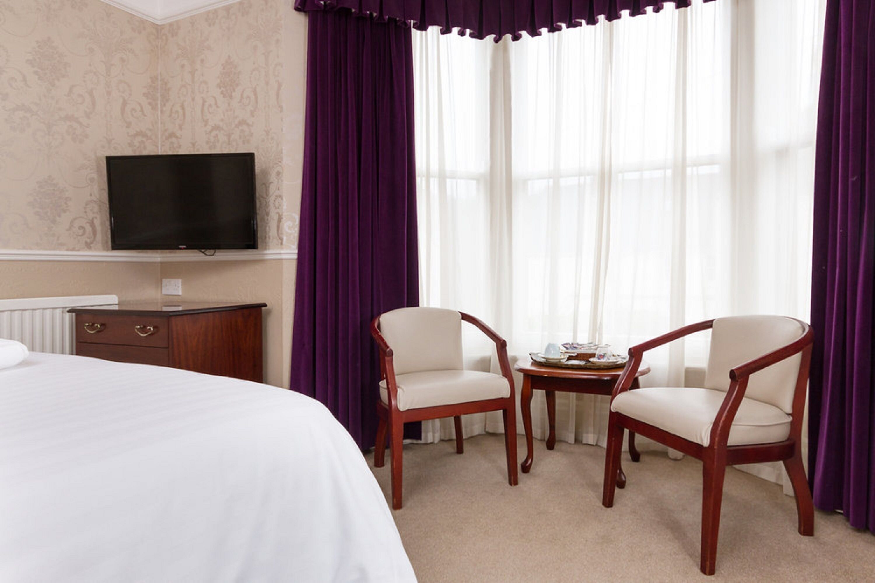 Habitación de hotel con cama, televisión, dos sillas, una mesita y ventana con cortinas.