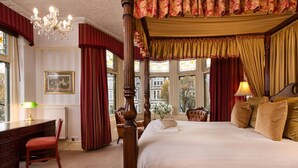 A four-poster bed with a canopy, a desk with a lamp, a chair, and a view of the city through large windows. - Lincoln House Hotel (Cardiff)