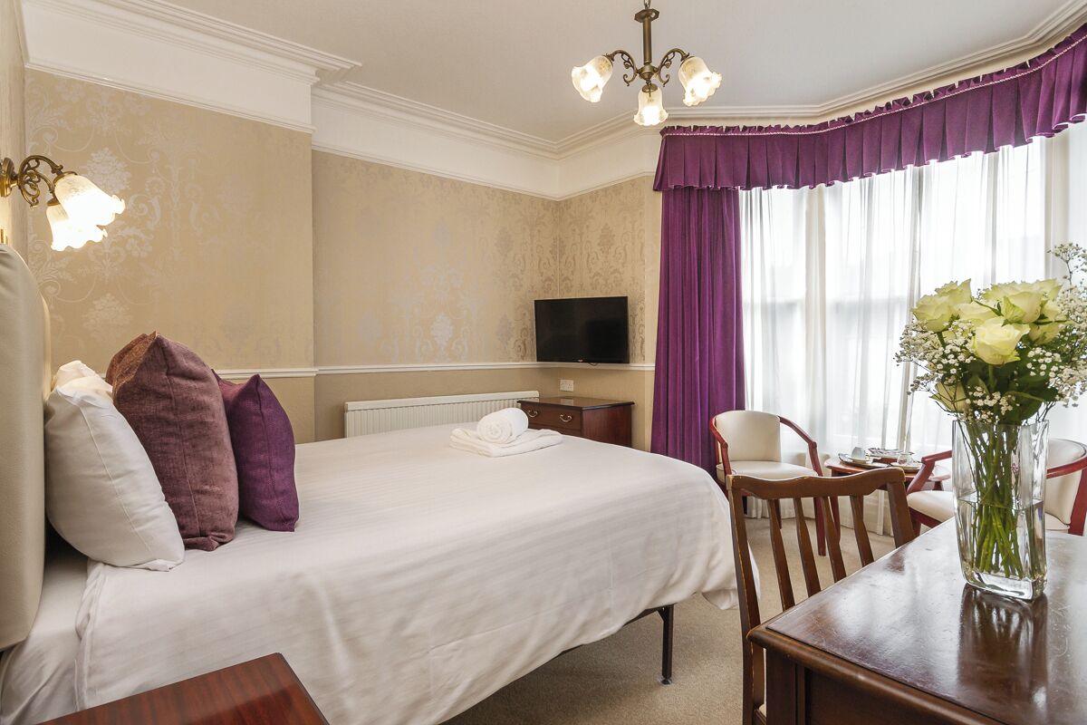 A hotel room with a large bed, a television, a dining table, and a vase with flowers.