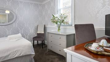 Ett hotellrum med en säng, en byrå, en stol, ett skrivbord med ett bricka med te och en fönster med persienner.