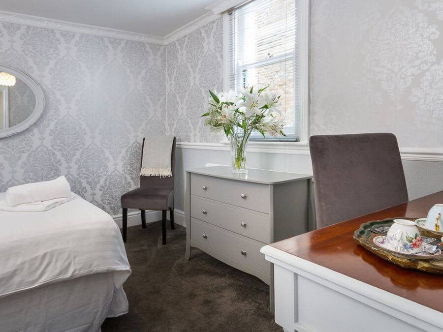 Une chambre d’hôtel comprenant un lit, une commode, une chaise, un bureau avec un plateau contenant du matériel pour préparer du thé, et une fenêtre avec des stores.