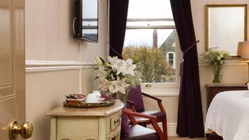 A hotel room with a bed, a desk with a vase of flowers, a chair, a television mounted on the wall, and a window with curtains.