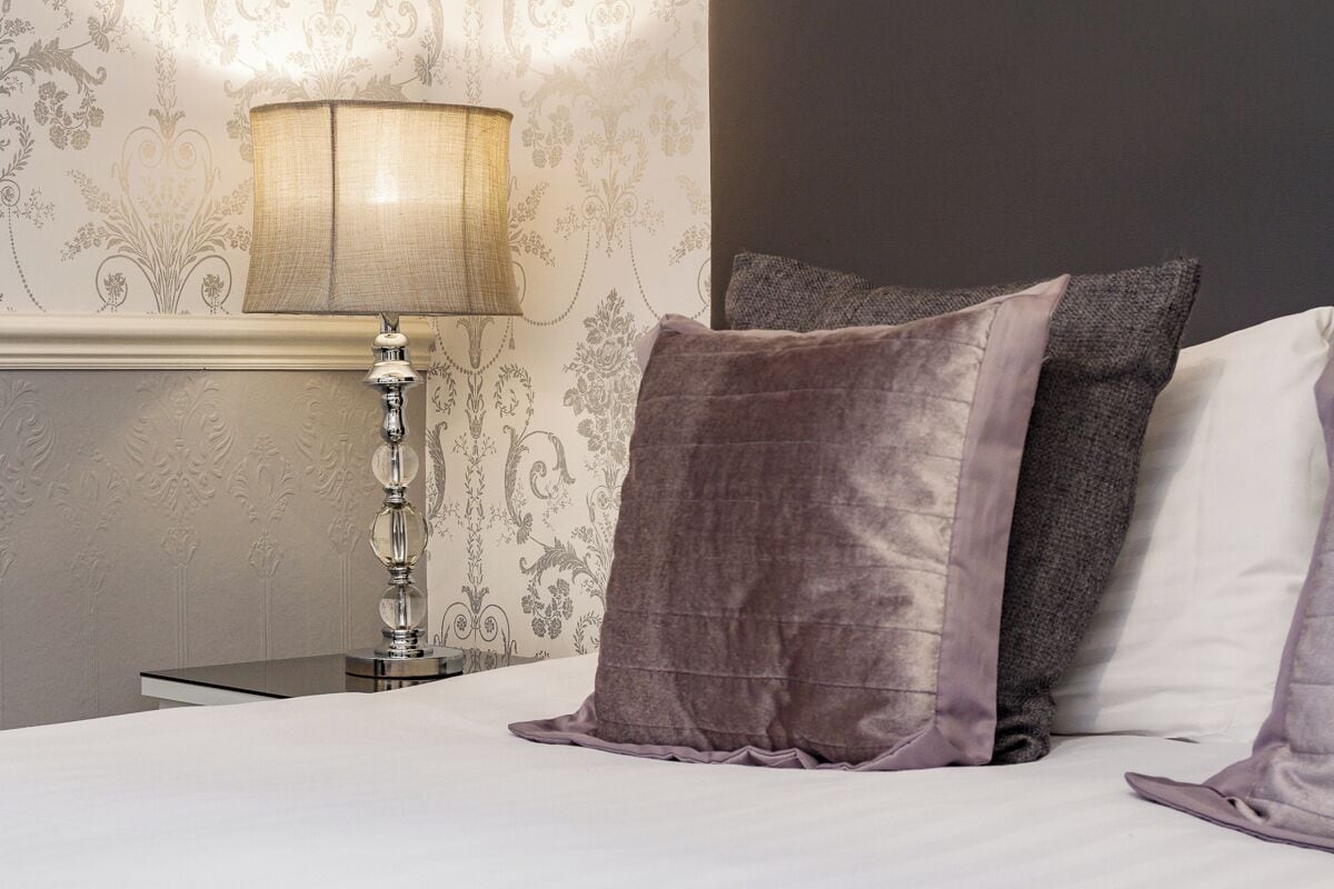 A neatly made bed with a white and gray pillow, a textured lamp, and a wall with floral wallpaper.