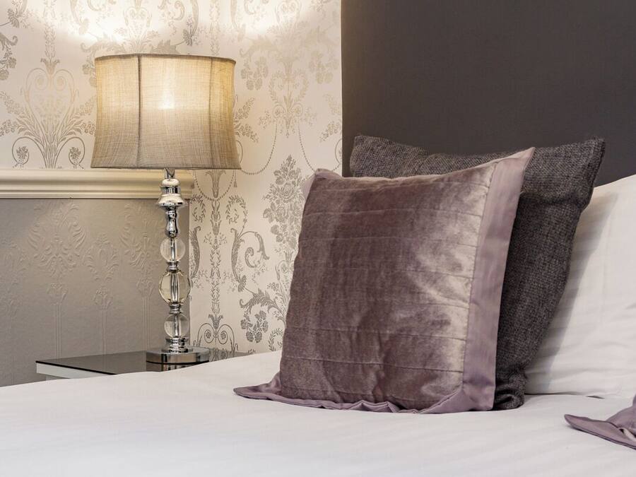 A neatly made bed with a white and gray pillow, a textured lamp, and a wall with floral wallpaper.