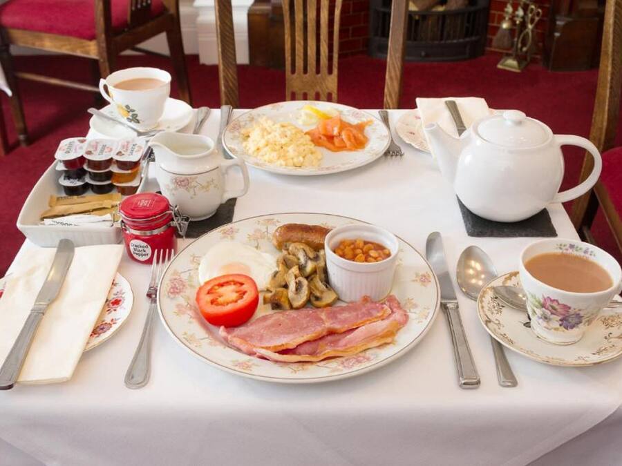 Mesa de café da manhã com pratos de ovos, bacon, cogumelos e feijão, além de xícaras de chá e café.