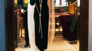 Una mujer con un vestido negro, velo y collar de perlas, de pie frente a un espejo.