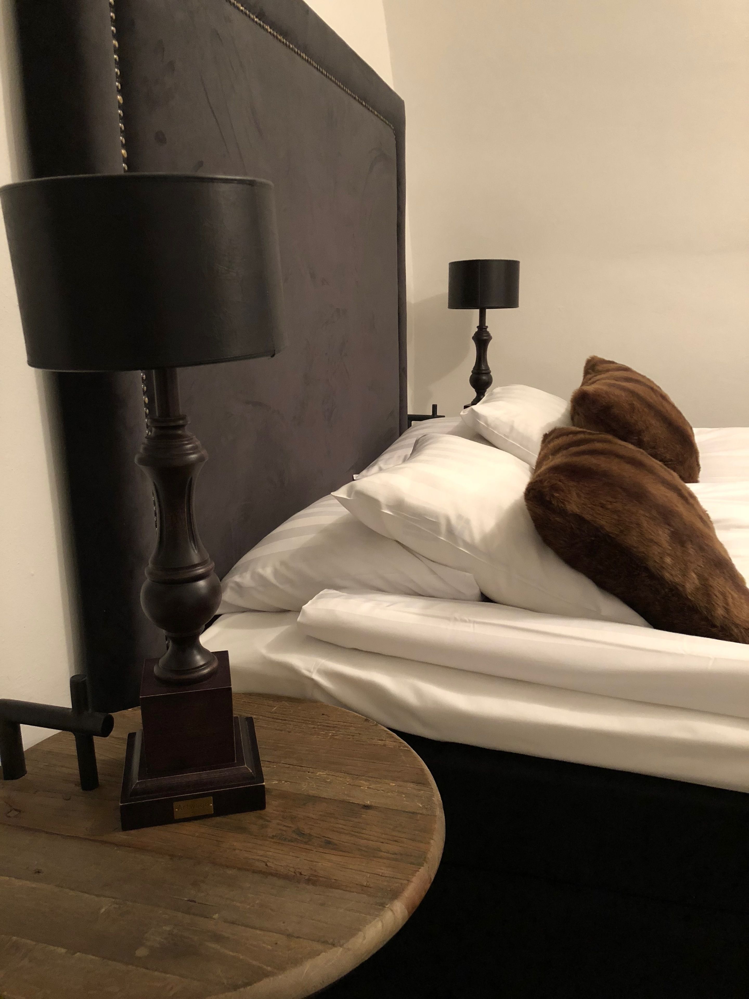 Uma cama bem arrumada com lençóis brancos e duas almofadas castanhas, uma mesa de cabeceira de madeira escura com abajur, e uma mesa de madeira redonda.