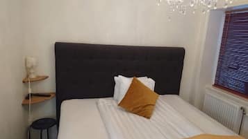 Un dormitorio con cabecera oscura, colcha blanca y almohada marrón. Se observa un pequeño estante de madera con un objeto blanco encima, y un radiador en la esquina.