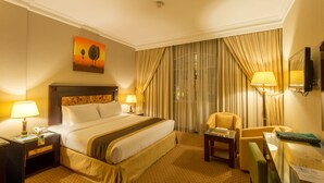 A hotel room with a large bed, two bedside lamps, a chair, a desk, and a painting on the wall. -  Caesar Hotel (Muscat)