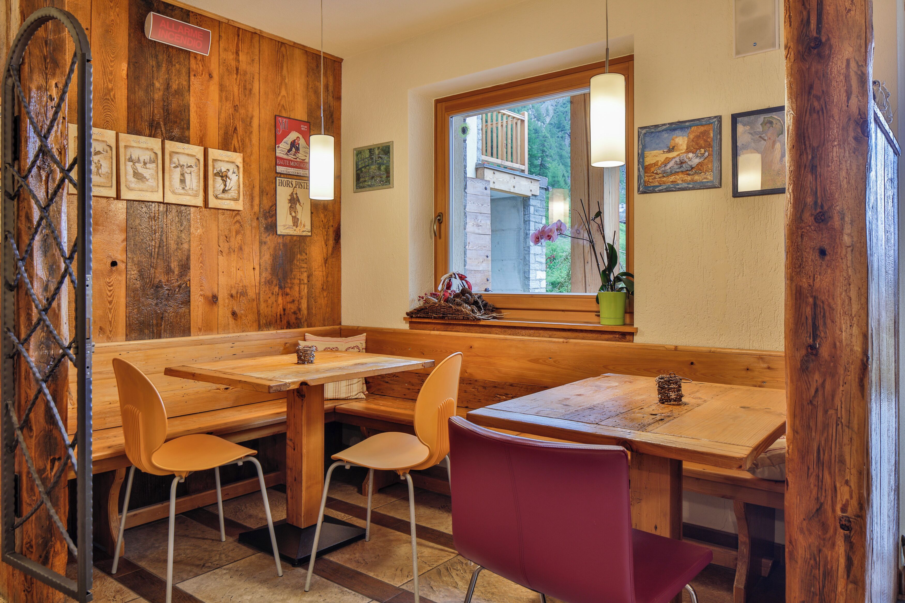 A rustic wooden interior with wooden benches and tables, framed pictures on the wall, and a window with a view of greenery outside.
