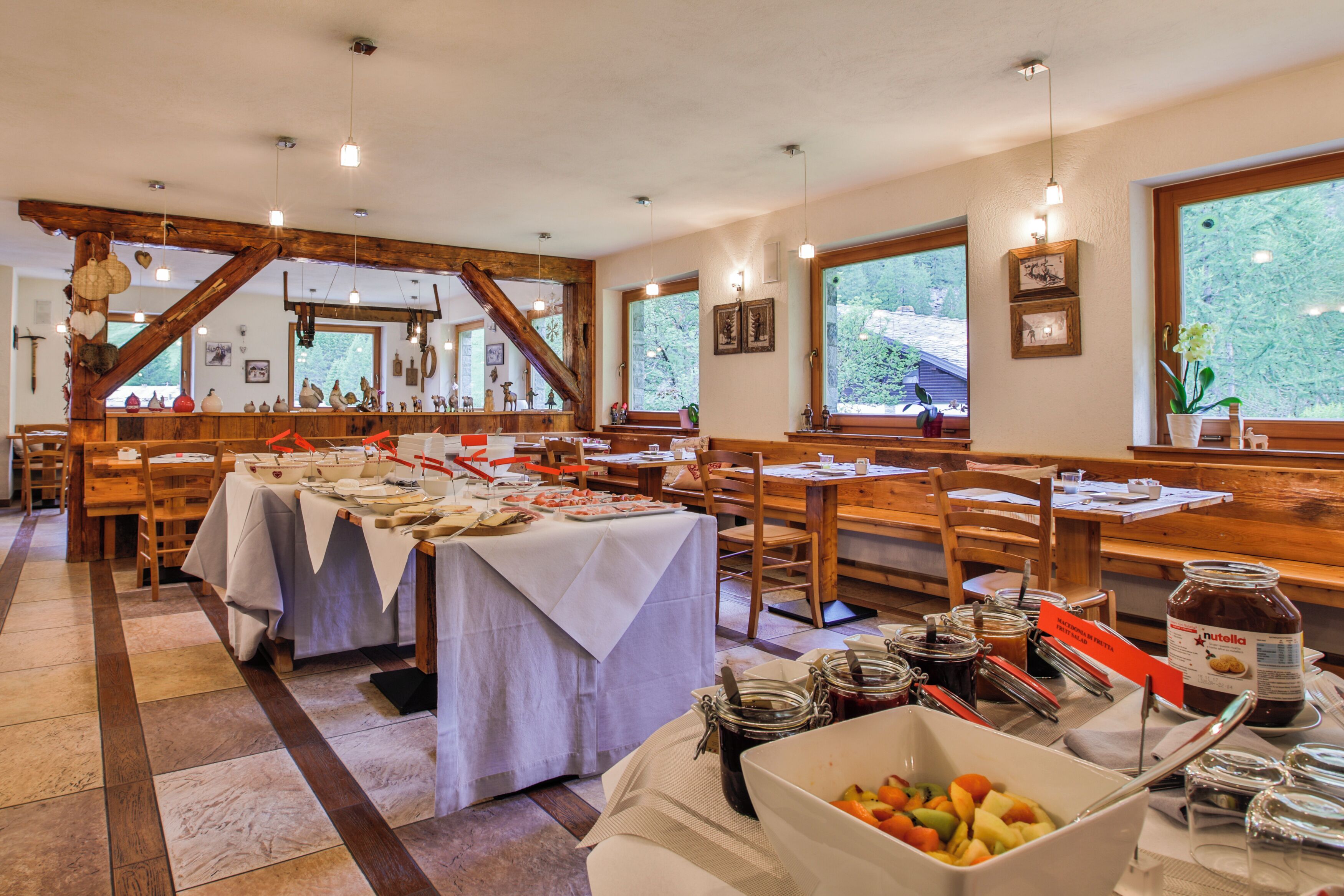A dining area with wooden tables and benches, a buffet setup with various food items, and a large window offering a view of greenery outside.