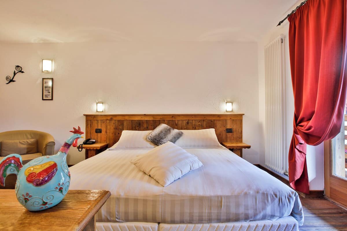 A hotel room with a wooden headboard, two bedside lamps, a bed with white linens, a red curtain, a decorative ceramic rooster, and a small table with a vase.