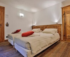 Un dormitorio con cabecera de madera, dos camas con ropa de cama color crema, una almohada roja y una pequeña lámpara de pared.