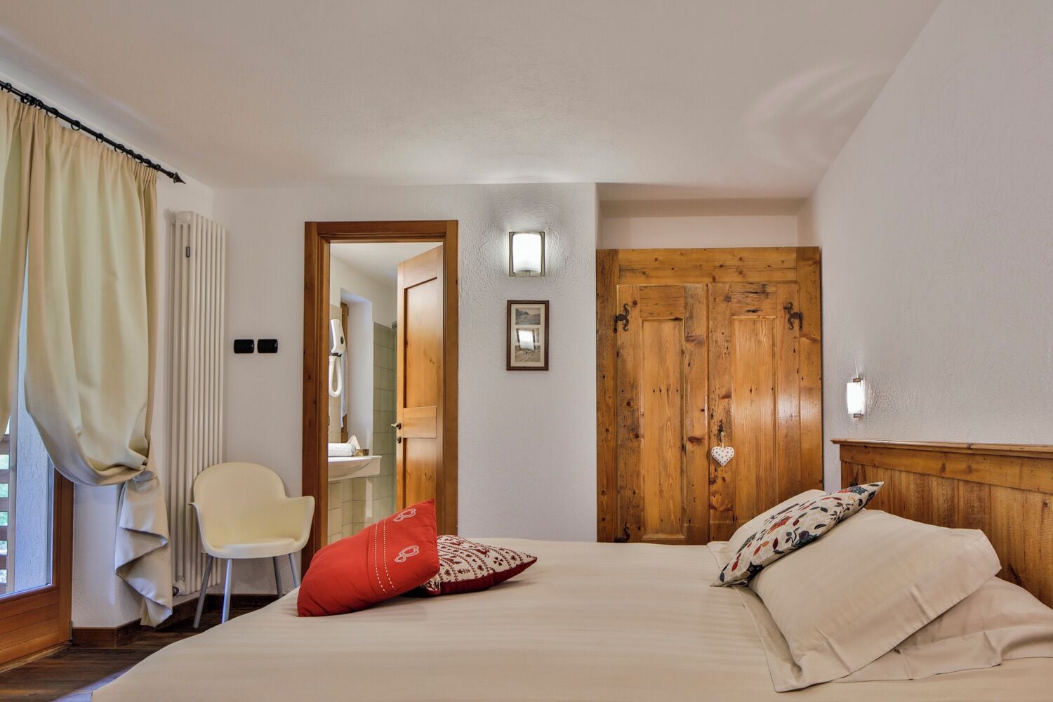 Quarto com cama, uma cadeira, guarda-roupa de madeira e banheiro visível por uma porta aberta.