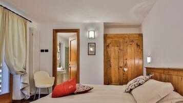 Quarto com cama, uma cadeira, guarda-roupa de madeira e banheiro visível por uma porta aberta.