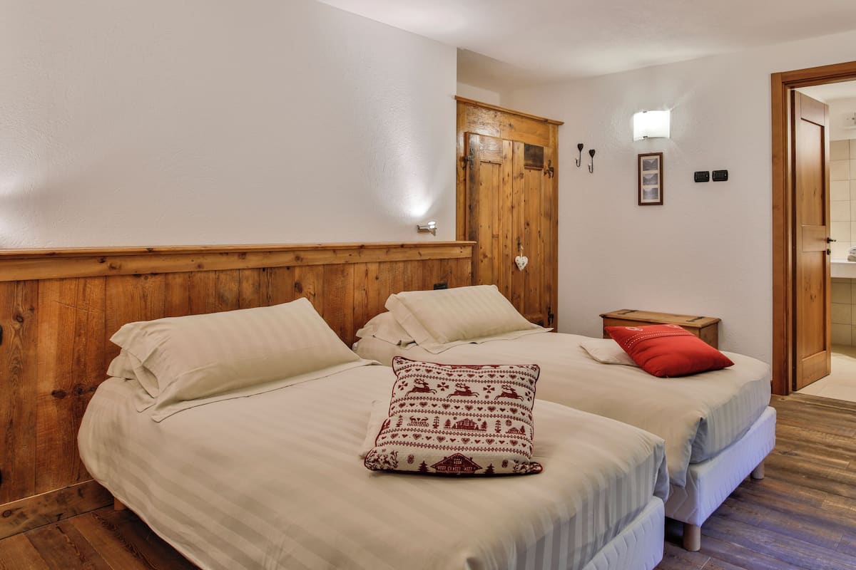A bedroom with two beds, wooden headboards, a wooden wardrobe, a bedside table with a red pillow, and a wall-mounted light fixture.