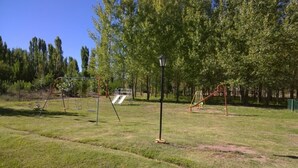 A playground with swings, slides, and a lamp post in a grassy area with trees in the background. - Cabañas Uspallata (Uspallata)