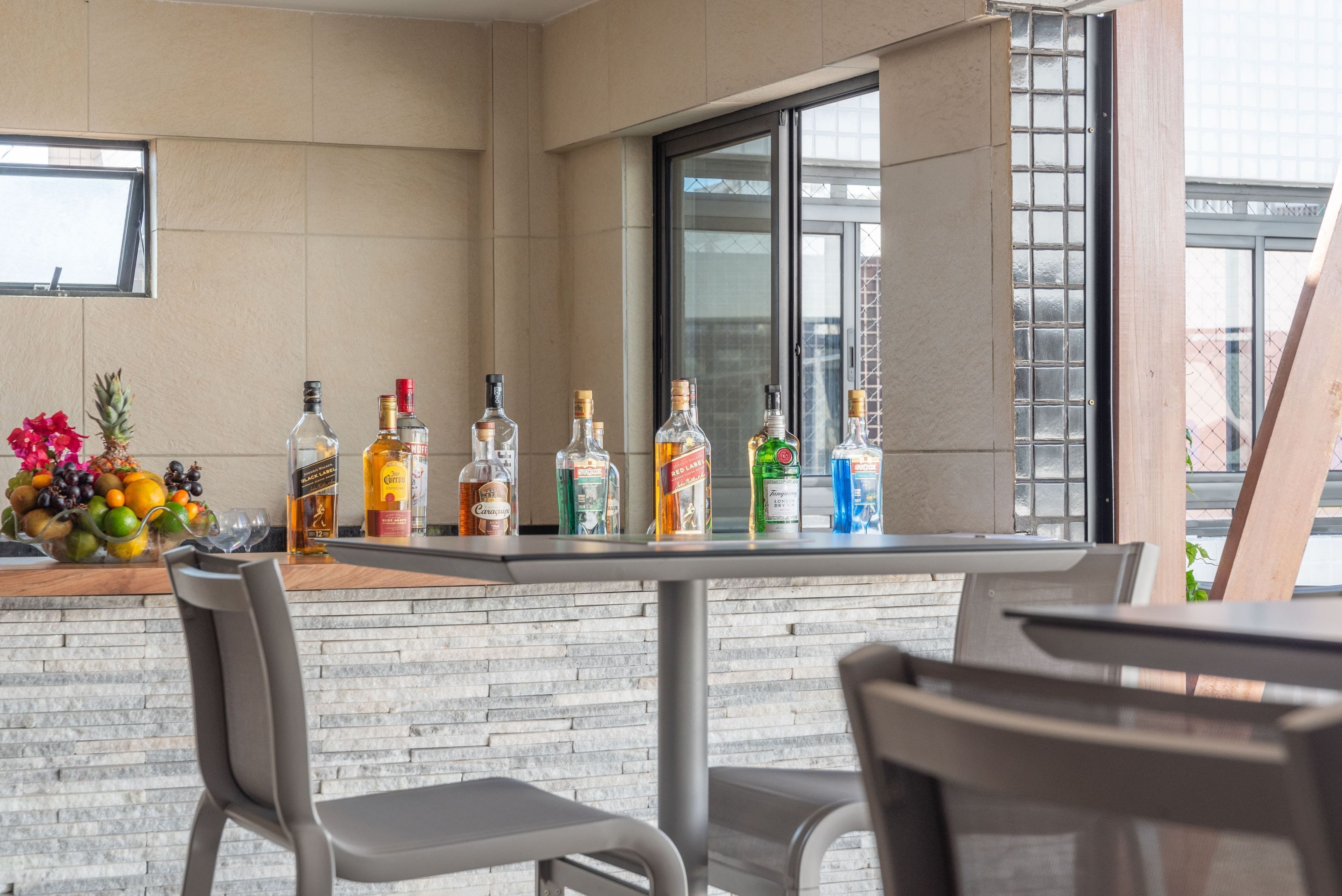 Zona de bar con mostrador que exhibe varias botellas, un frutero y dos sillas.