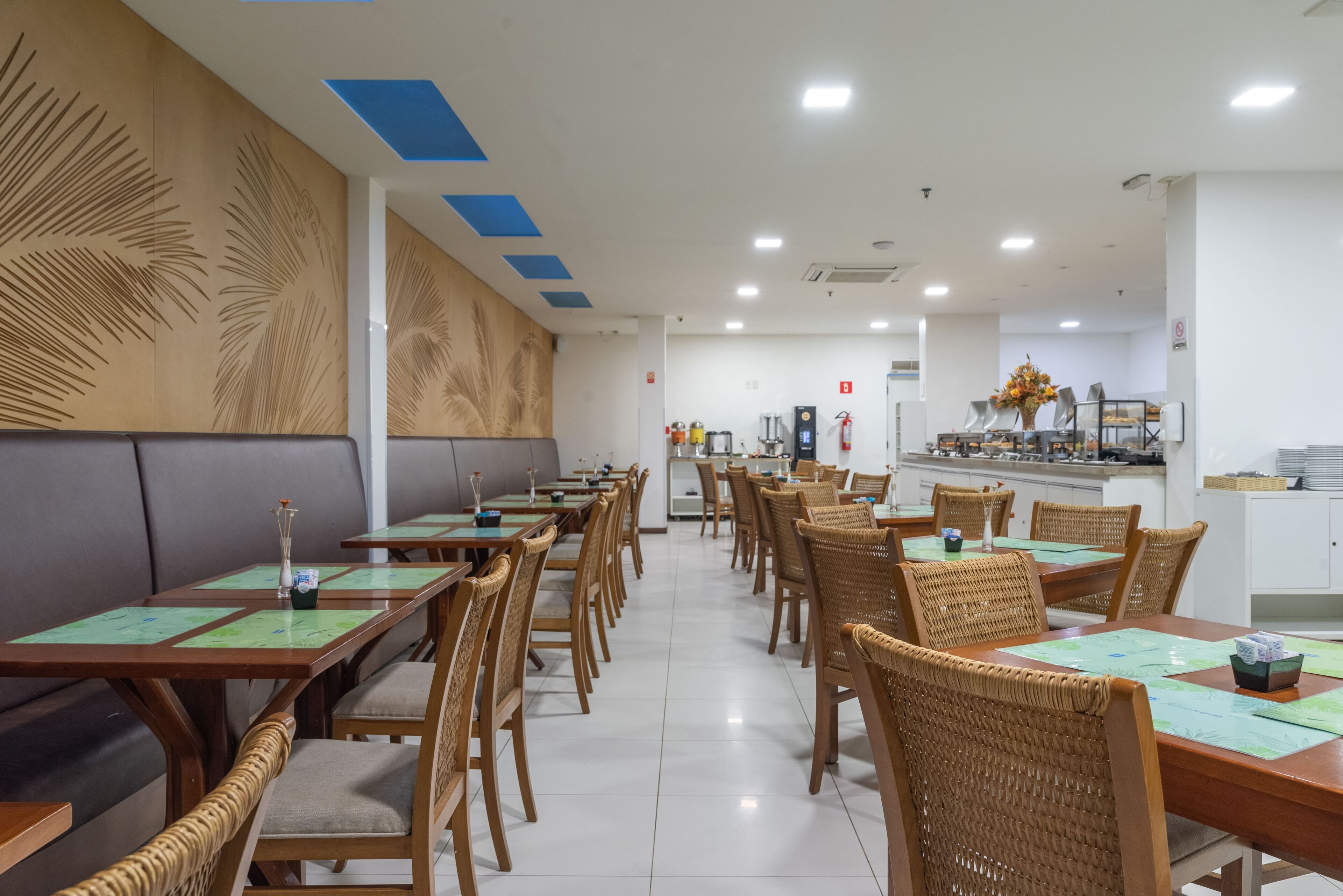 Ein Restaurant mit langen Holztischen, Korbstühlen, einer Theke mit Registrierkasse und einer Wand mit Palmblatt-Mustern.