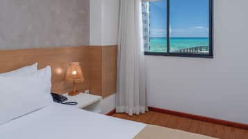 A hotel room with a bed, a bedside table with a lamp, a phone, a window with a view of the ocean, and a wooden floor.
