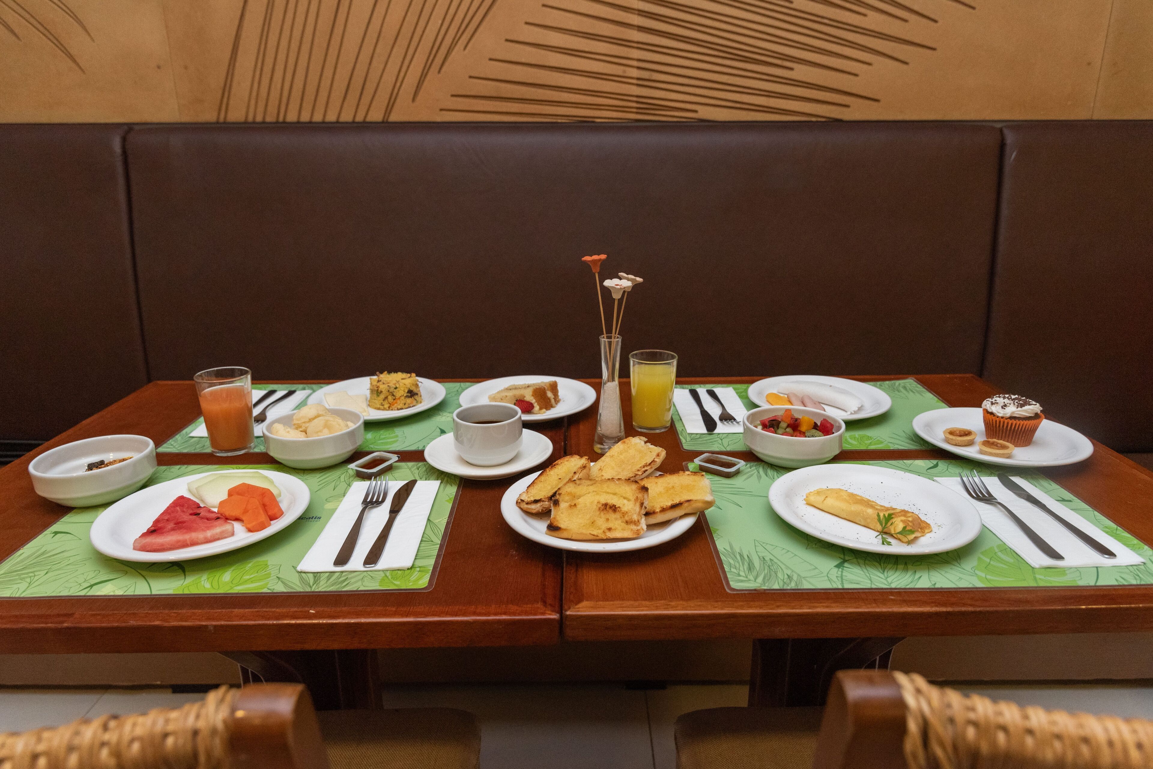 A dining table set with breakfast items including toast, fruit, and a muffin, with a green placemat and a decorative vase.