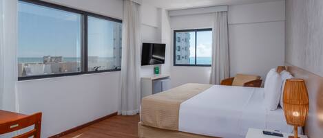 A hotel room with a bed, a TV, a desk, a chair, and large windows offering a view of the sea.