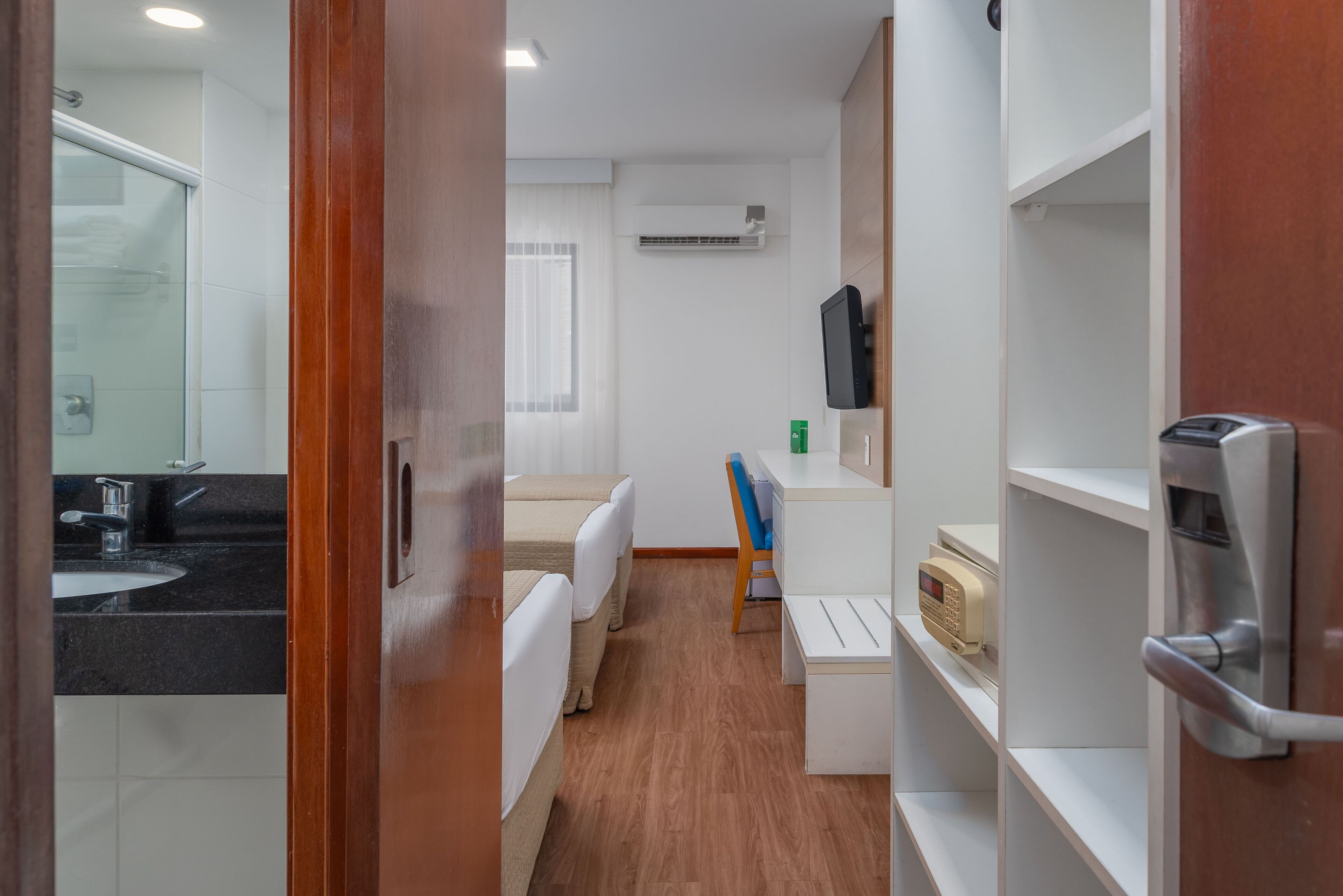 A hotel room with a wooden floor, a white sink, a bathtub, a desk with a chair, and a TV mounted on the wall.
