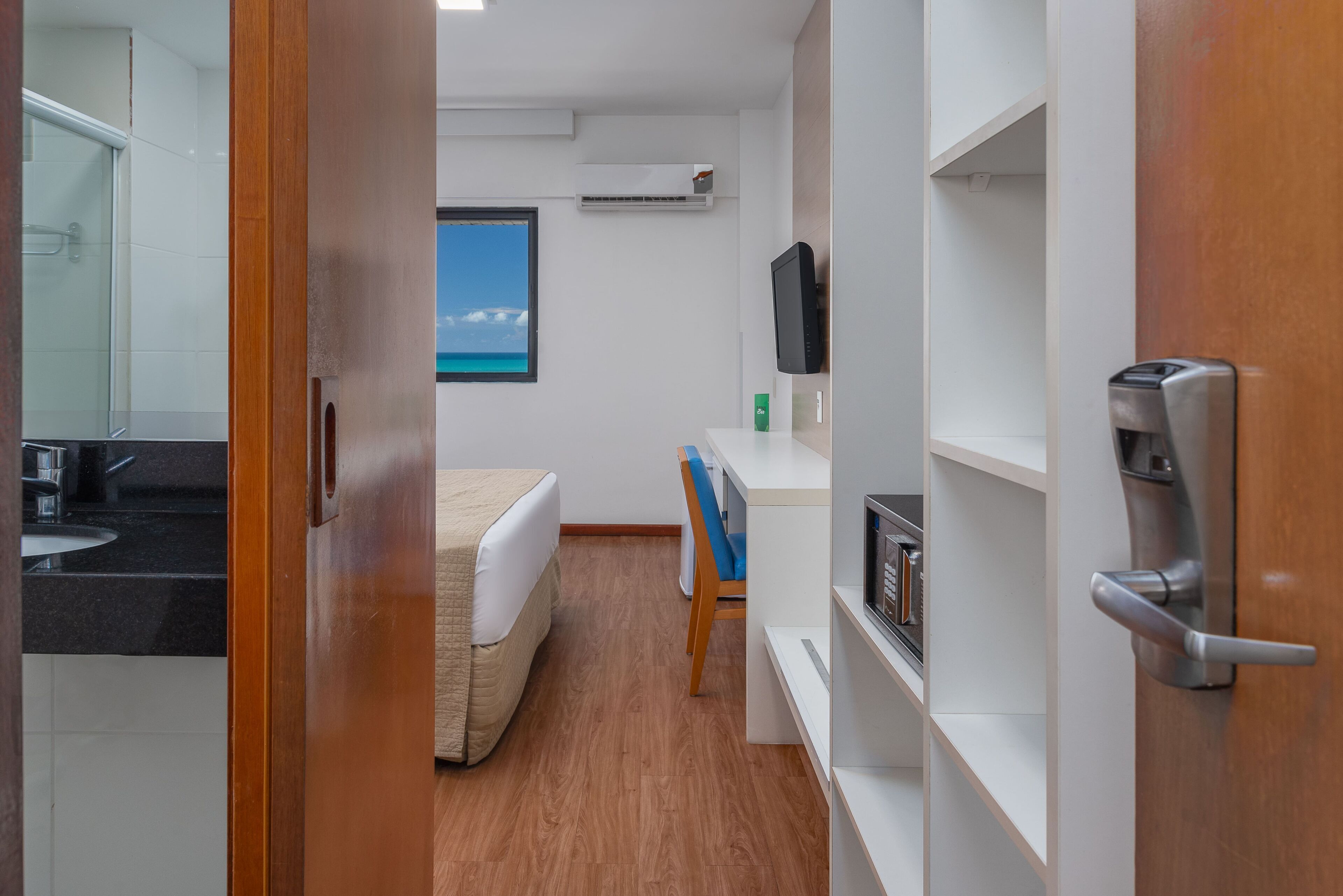 A hotel room with a bed, a desk, a chair, a TV, a shelf with books, and a coffee machine.