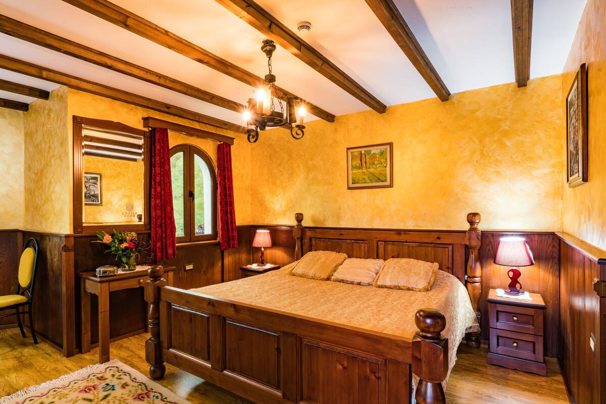 A wooden four-poster bed with a nightstand and lamp, a desk with a chair, and a window with curtains.