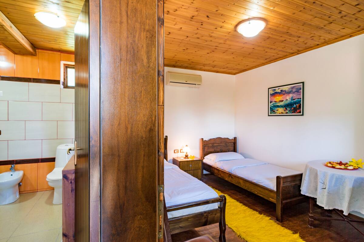 A room with wooden ceiling and walls, a bed, a bedside table with a lamp, a small table with a fruit plate, and a painting on the wall.