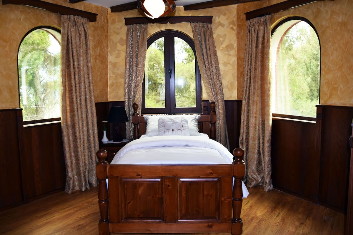 A wooden four-poster bed with a white duvet, flanked by two arched windows with curtains, in a room with wooden paneling and a ceiling light fixture.